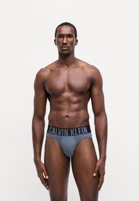 Sivi pamučni gaćice sa crnim pojasom na kojem je logo "CALVIN KLEIN". Model se čvrsto prianja uz tijelo, ističući oblik. Teksturisani materijal.