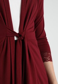 Bordowy kardigan z wiązanym w talii paskiem, wykonany z miękkiego, elastycznego materiału, z koronkowymi dodatkami przy mankietach rękawów. Gładka faktura i luźny krój.