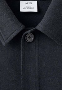 Camicia giacca nera in misto lana con colletto a punta, chiusura a un bottone e texture liscia. L'etichetta indica la taglia e l'origine.