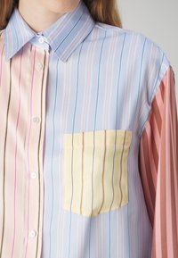 Meerkleurig gestreept shirt met een lichtblauwe basis, roze mouwen, een gele zak en dunne verticale lijnen in roze, blauw en bruin.