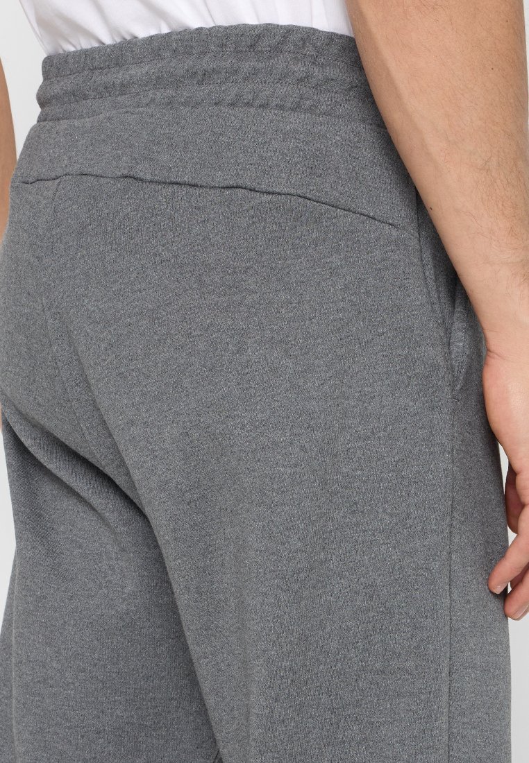Uomo che indossa pantaloni sportivi grigi a maglia e una maglietta bianca, mostra un primo piano dell'elastico in vita e una mano che riposa di lato.