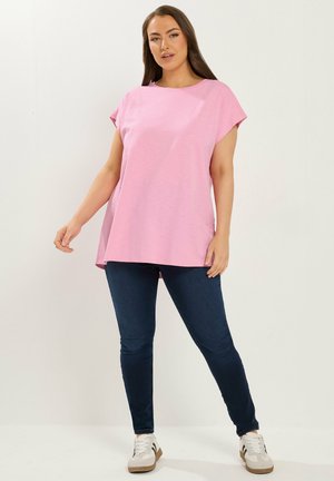 Kobieta w luźnym różowym topie z krótkim rękawem, ciemnoniebieskich dopasowanych dżinsach i białych trampkach, stojąca na tle jednolitego jasnego tła.