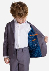 Sivi suknjič z izrezanim klopom, na en gumb in modrim vzorčastim podlogom. Ima belo srajco spodaj in siva hlače.