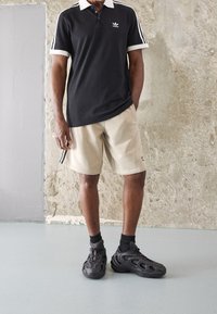 Man som bär en svart Adidas-polo, beige Adidas-shorts, svarta strumpor och svarta träningsskor, står på en grå golv mot en texturerad vägg.