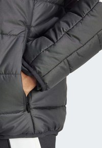 Svart pufferjakke med kviltet design, med en glidelåslomme på siden og ribbestrikkede mansjetter. Glatt stoff med en svak glans.