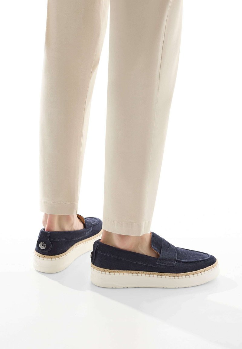 Marine slip-on schoenen met een gestructureerde bovenkant, witte rubberen zool en decoratieve stiksels langs de rand. Gecombineerd met lichtgekleurde broeken.