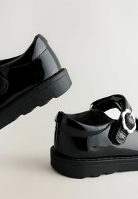 Chaussures vernies noires à bout fermé, avec sangle réglable et détail de boucle ronde, dotées d'une semelle texturée noire et d'une finition lisse.