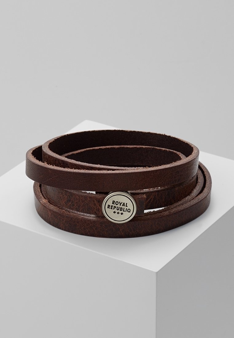 Bracelet en cuir marron foncé, enroulé, avec un fermoir métallique rond gravé de "ROYAL REPUBLIQ" sur un socle blanc.