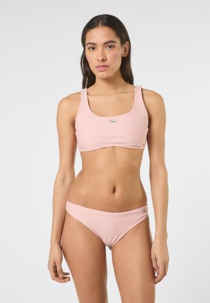 Kvinde med langt mørkt hår iført en lyserød Fila sports-bh og matchende bikinitrusser, stående mod en ensfarvet baggrund.