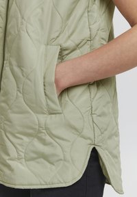 Quilted Weste in Hellgrün, mit einem Reißverschluss und seitlichen Taschen. Weiche, glatte Textur mit wellenförmiger Steppung und abgerundetem Saum.