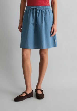 Femme portant une jupe bleue à taille élastique avec cordon, un top rouge et des ballerines marron Mary Jane, debout devant un fond gris uni.