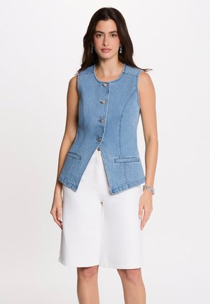 Frau trägt ärmellose blaue Jeansweste mit Knöpfen und weiße kniehohe Shorts, steht vor einem einfarbigen hellen Hintergrund.