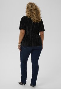 Donna con capelli ricci che indossa un top corto in velluto nero, jeans blu scuri, tacchi neri e un braccialetto di perle, in piedi di spalle.