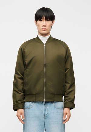 Young man wearing an olive green bomber jacket over a white turtleneck and light blue jeans, standing against a plain white background.