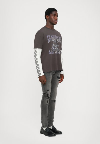 Grijze graphic t-shirt met witte lange mouwen met een ruitjespatroon, versleten grijze jeans en zwarte loafers. Eenvoudig ontwerp en casual pasvorm.