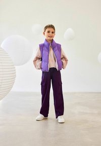 Lilla puffer vest over en lys rosa sweatshirt med oransje detaljer, kombinert med løstsittende lilac cargo bukser og hvite joggesko.