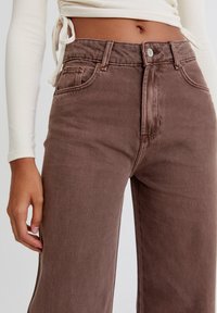 Ruske korkeavyötäröiset farkut, joissa on leveät lahkeet, etutaskut ja metallinen nappikiinnitys. Tasainen kangas, jossa on hienovaraisia ompeluyksityiskohtia.