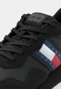 Schwarzer Sneaker mit einem glatten Lederobermaterial, ausgestattet mit einfarbigen Schnürsenkeln und einem mehrfarbigen Seitenstreifen in Marineblau, Rot und Weiß. Logo auf der Zunge.