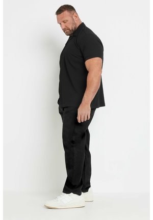 Mann mit kurzen Haaren und Bart steht seitlich, trägt ein schwarzes Kurzarmhemd, schwarze Hose und weiße Sneaker vor einem schlichten Hintergrund.