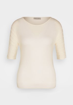 Haut à manches courtes de couleur crème en tissu doux, avec un col rond et une texture subtile sur les manches. Étiquette visible au niveau du col.