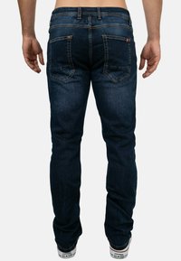 Jeans in denim blu scuro con una vestibilità slim, due tasche posteriori e sottili dettagli di cucitura. Il tessuto sembra liscio con una leggera consistenza.