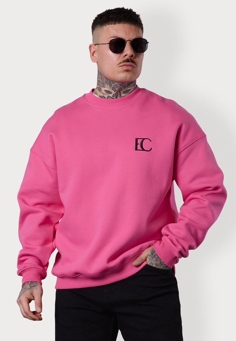 Sweatshirt en tissu rose vif avec des poignets et un ourlet côtelés, arborant un petit logo noir sur la poitrine. Le modèle porte un jean foncé et des lunettes de soleil.