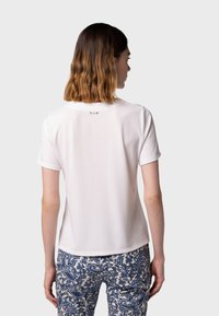 Bílý krátký rukáv top s kulatým výstřihem, který na zadní straně obsahuje jemnýDetail. Nosi se s vzorovanými modro-krémovými kalhotami. Hladká textura.