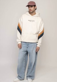 Witte hoodie met blauwe en oranje strepen, voorzien van een voorzak en logo. Gecombineerd met lichtblauwe wijde jeans en witte sneakers.