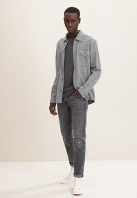Camisa de botones gris sobre una camiseta gris oscuro, combinada con jeans gris desgastado y zapatillas blancas. Cuenta con dos bolsillos en el pecho y un corte relajado.