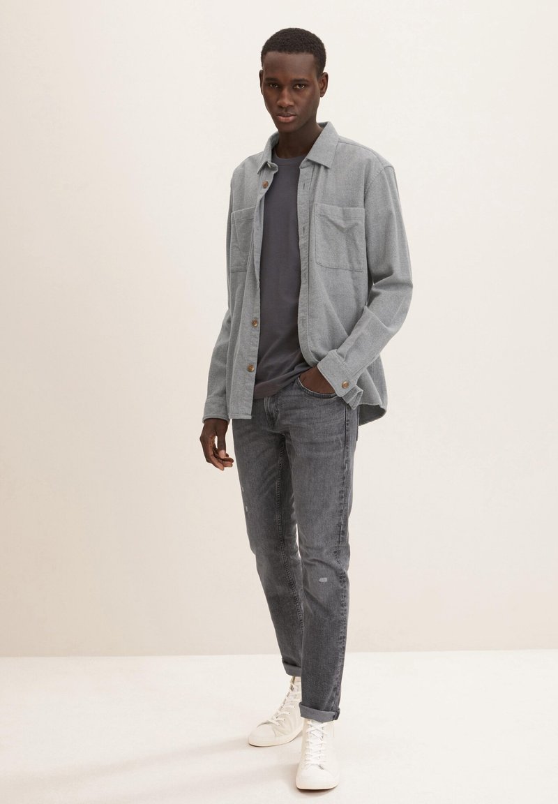 Camisa de botones gris sobre una camiseta gris oscuro, combinada con jeans gris desgastado y zapatillas blancas. Cuenta con dos bolsillos en el pecho y un corte relajado.
