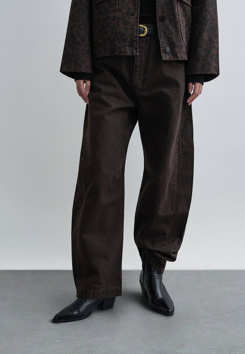 Calças castanhas de perna larga em tecido de ganga espessa, apresentando um corte descontraído e detalhes subtis de costura. Combinadas com botas pretas de salto apontadas.