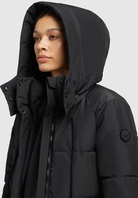 Black puffer jacket with a large hood, zip closure, and snap buttons. Features a logo patch on the sleeve and quilted texture.
