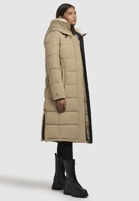 Sieviete ar garu beigu krāsas mīksto kažoku ar kapuci un melnām masīvajām šņorējamām zābakiem, stāv sānis uz vienveida fona.