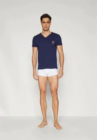 T-shirt bleu marine à col en V, manches courtes, tissu doux ; slips blancs. Accent de logo subtil sur la poitrine, modèle se tenant devant un fond neutre.