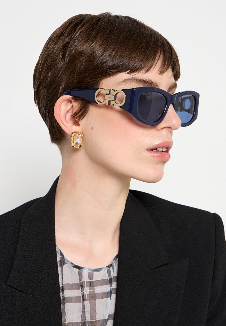 Frau mit kurzen braunen Haaren, die marineblaue Cat-Eye-Sonnenbrillen mit goldenen Akzenten, goldene Edelsteinohrringe, einen schwarzen Blazer und eine karierte Bluse trägt.