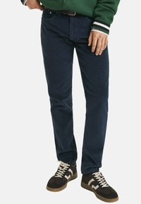 Marineblauwe corduroy broek met een rechte pasvorm, voorzien van een klassieke vijfzakkenontwerp, en gecombineerd met donkere sneakers en een groen zip-up jack.