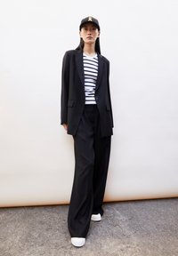 Zwarte blazer over een wit met zwart gestreept shirt, gecombineerd met brede zwarte broek en witte sneakers, aangevuld met een zwarte pet met een gele accent.