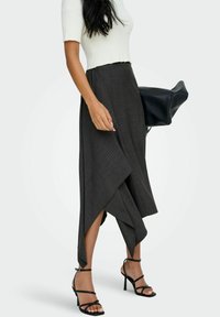 Vrouw in een witte ribbelige top met korte mouwen, een asymmetrische rok met donkere streepjes, zwarte sandalen met bandjes en een grote zwarte handtas.