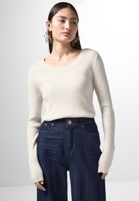 Geribbelde gebreide trui in beige met een brede halslijn, gecombineerd met donkerblauwe wijde broekspijpen. Opvallende lange mouwen en een aansluitende structuur.