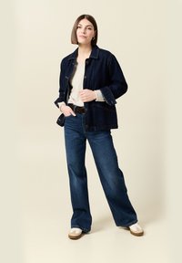 Vrouw die met haar handen in de zakken staat, gekleed in een donkerdenim jack, lichte trui, wijde broek en witte sneakers, op een beige achtergrond.