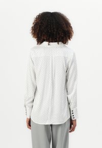 Person mit lockigem, dunklem Haar, der ein weißes Langarmshirt mit schwarzen Pünktchen und graue Hosen trägt, von hinten gezeigt.