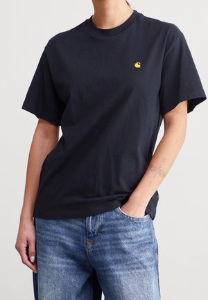 Person wearing a black short-sleeve t-shirt with a small yellow logo on the left chest, paired with blue jeans and a butterfly tattoo on the arm.