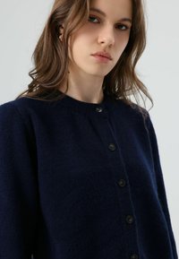 Marineblauwe cardigan van gestructureerde stof met een ronde halslijn en zes zwarte knopen aan de voorkant, met een eenvoudig, klassiek ontwerp.