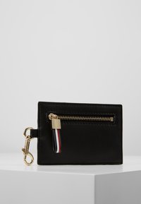 Porte-cartes en cuir noir avec une poche zippée dorée et un tire-zip tricolore, avec une fermeture métallique dorée sur le côté.