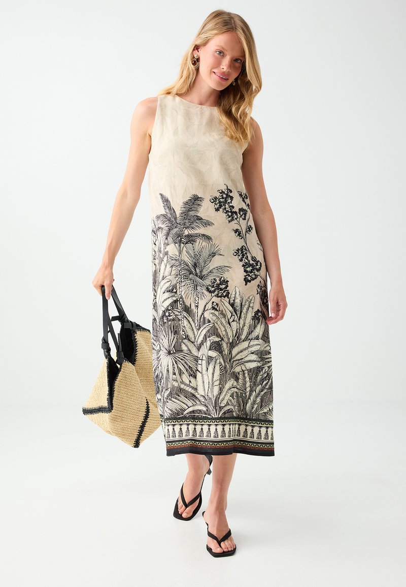 Femme portant une robe beige sans manches avec un imprimé botanique noir, tenant un sac tissé et portant des sandales noires, marchant vers l'avant.