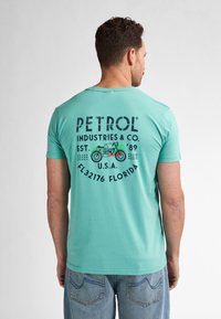 Tyrkysové bavlněné tričko s kulatým výstřihem, na zádech s velkým černým a zeleným grafickým potiskem obsahujícím text a ilustraci motocyklu.