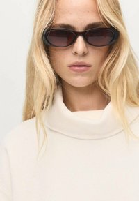 Occhiali da sole neri a forma ovale con lenti scure, indossati con un maglione a collo alto di colore crema. Texture liscia, design minimalista e colori neutri.