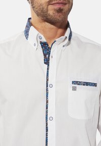 Weißes Hemd mit knopfbund, das einen gemusterten blau-braunen Kragen und Taschenbesatz aufweist. Es hat runde Knöpfe und eine einzige Brusttasche.