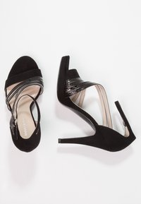 Černé sandály na vysokém podpatku s nubukovým svrškem, lesklými černými pásky a štíhlým jehlovým podpatkem. Mají otevřenou špičku a pásek kolem kotníku.