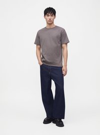 Jeune homme debout, une main dans la poche, portant un t-shirt gris à manches courtes, un jean large bleu foncé et des chaussures noires sur fond blanc.